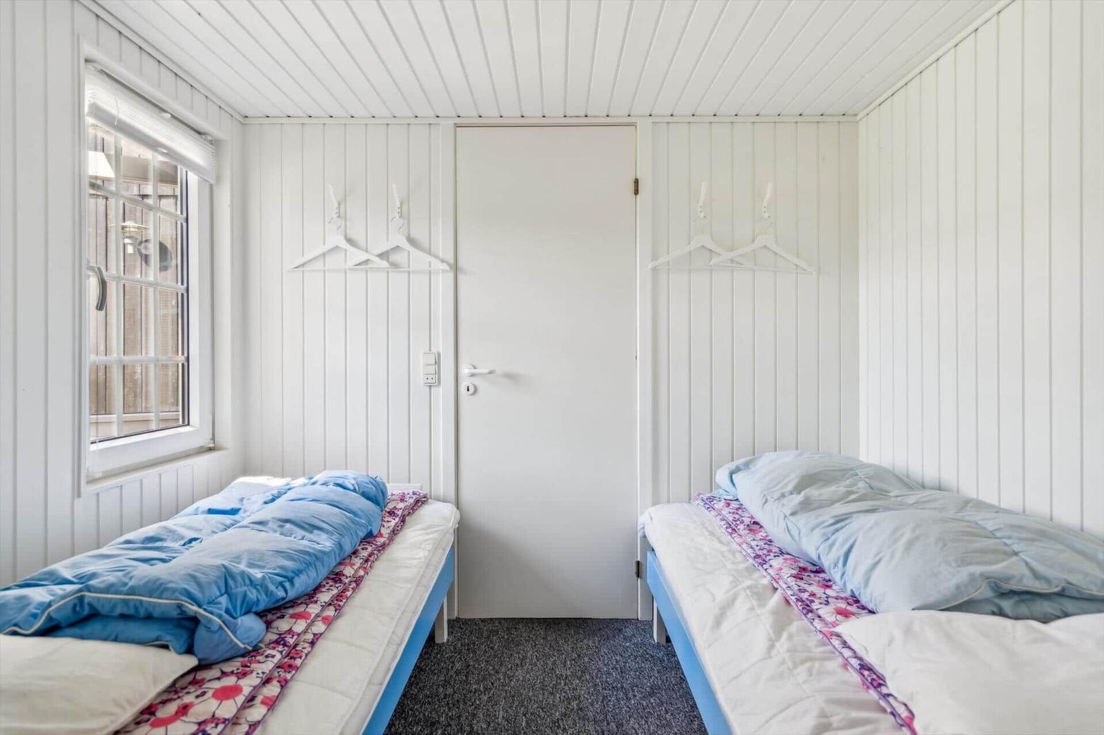 Zwei Betten mit blauen Decken und gemusterten Laken stehen in einem Raum mit weißen Wänden und einem Fenster.