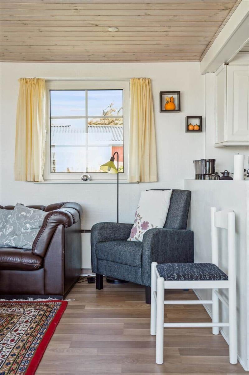 Wohnraum mit braunem Ledersofa, grauer Sessel, Holzboden und Fenster mit gelben Vorhängen.