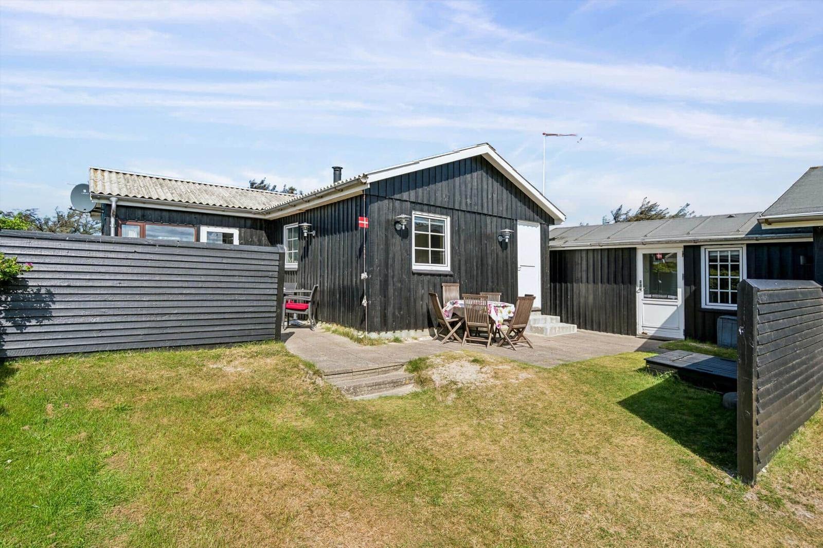 Ein schwarzes Holzhaus mit Terrasse und Garten. Es gibt einen Tisch mit Stühlen und eine Satellitenschüssel.