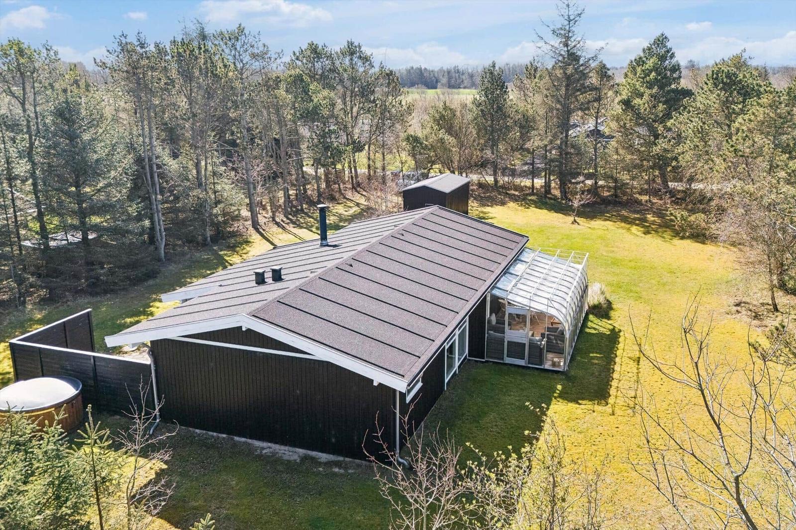 Modern wooden cabin with glass conservatory, surrounded by forest and lawn.