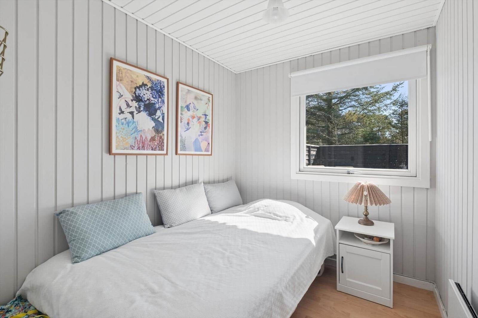 A bedroom with a bed, window, and nightstand. Two pictures hang on the walls.