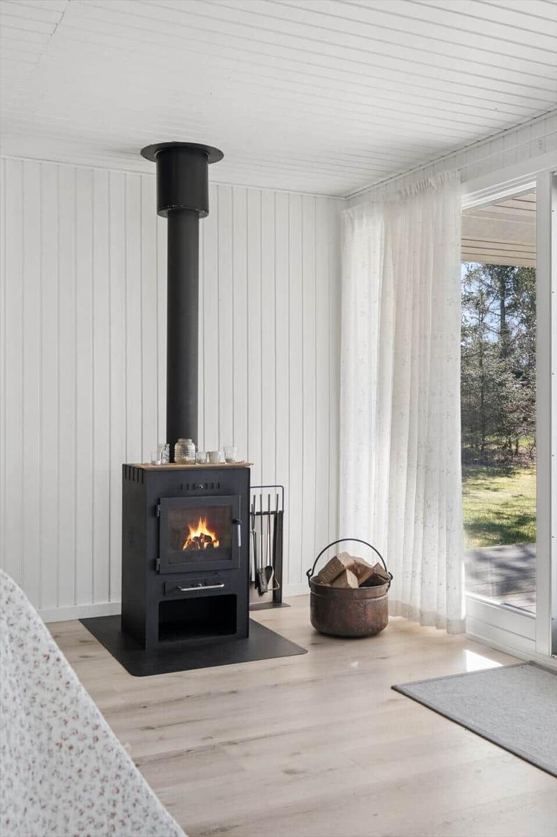 Black wood stove with fire, firewood basket, and view of green areas.