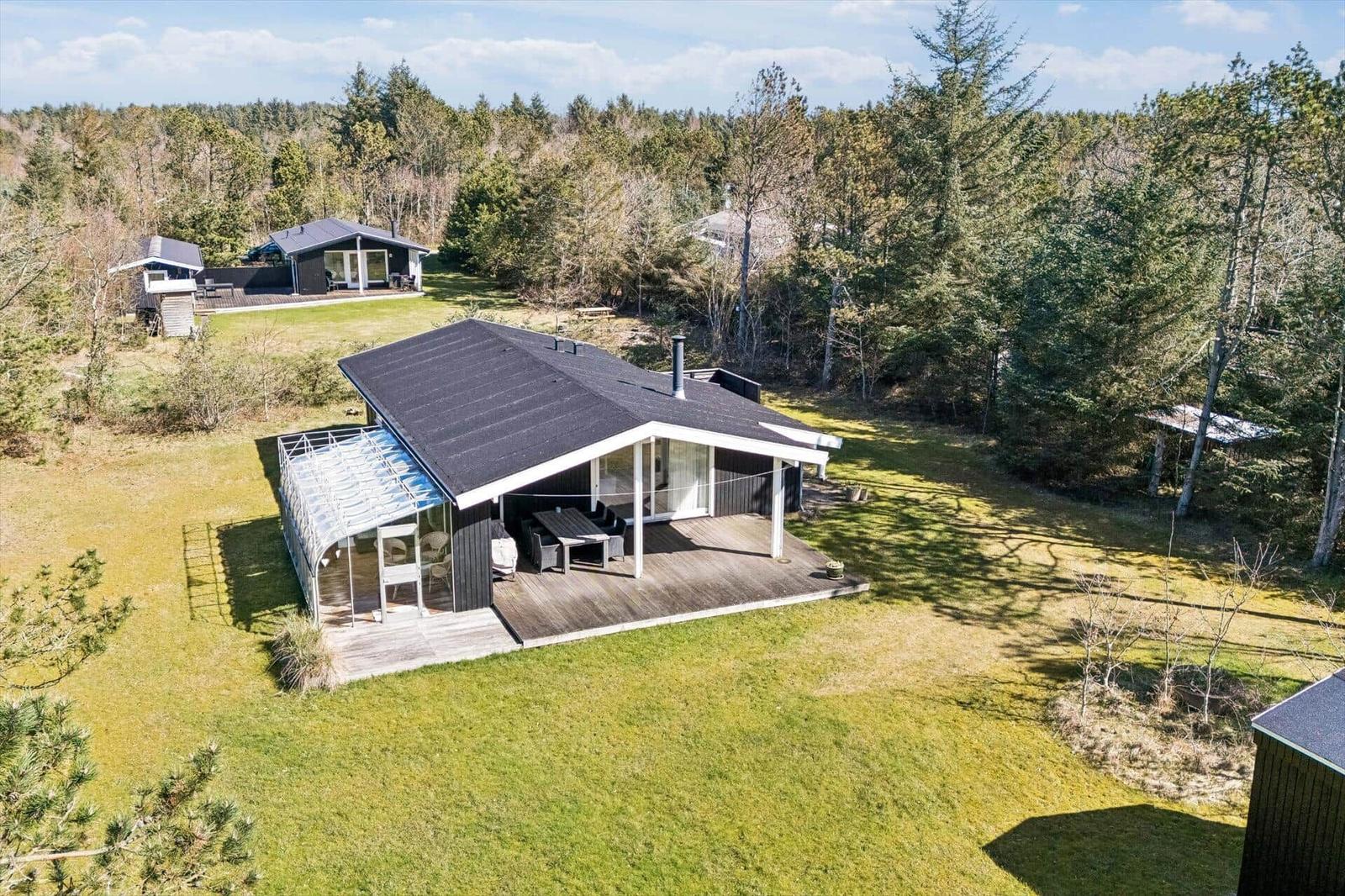 Modern wooden cabin with terrace and grill area in the forest. Green lawn and trees surround the house.