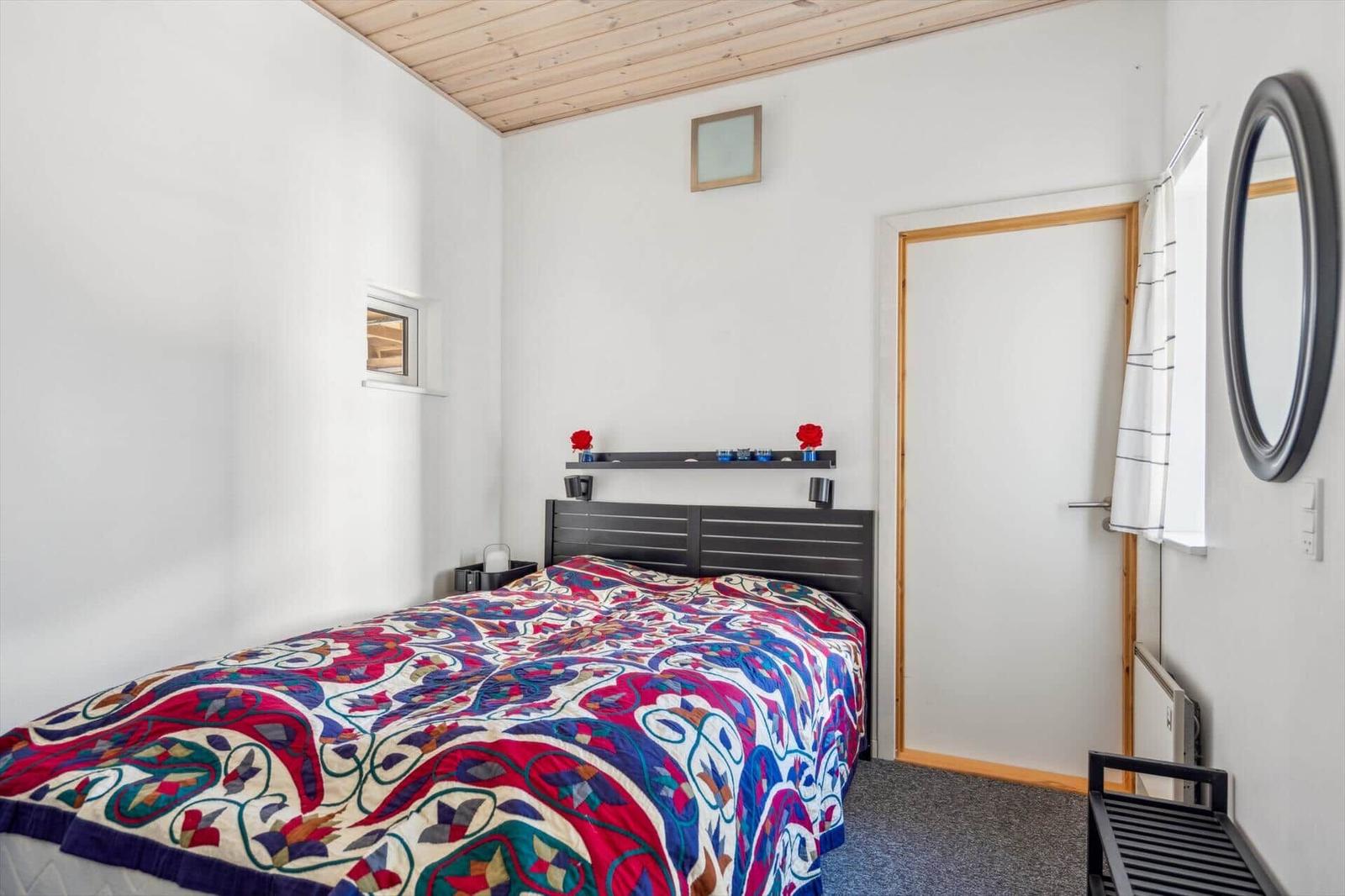 Schlafzimmer mit Bett, Kopfteil und Wandspiegel. Decke aus Holz.