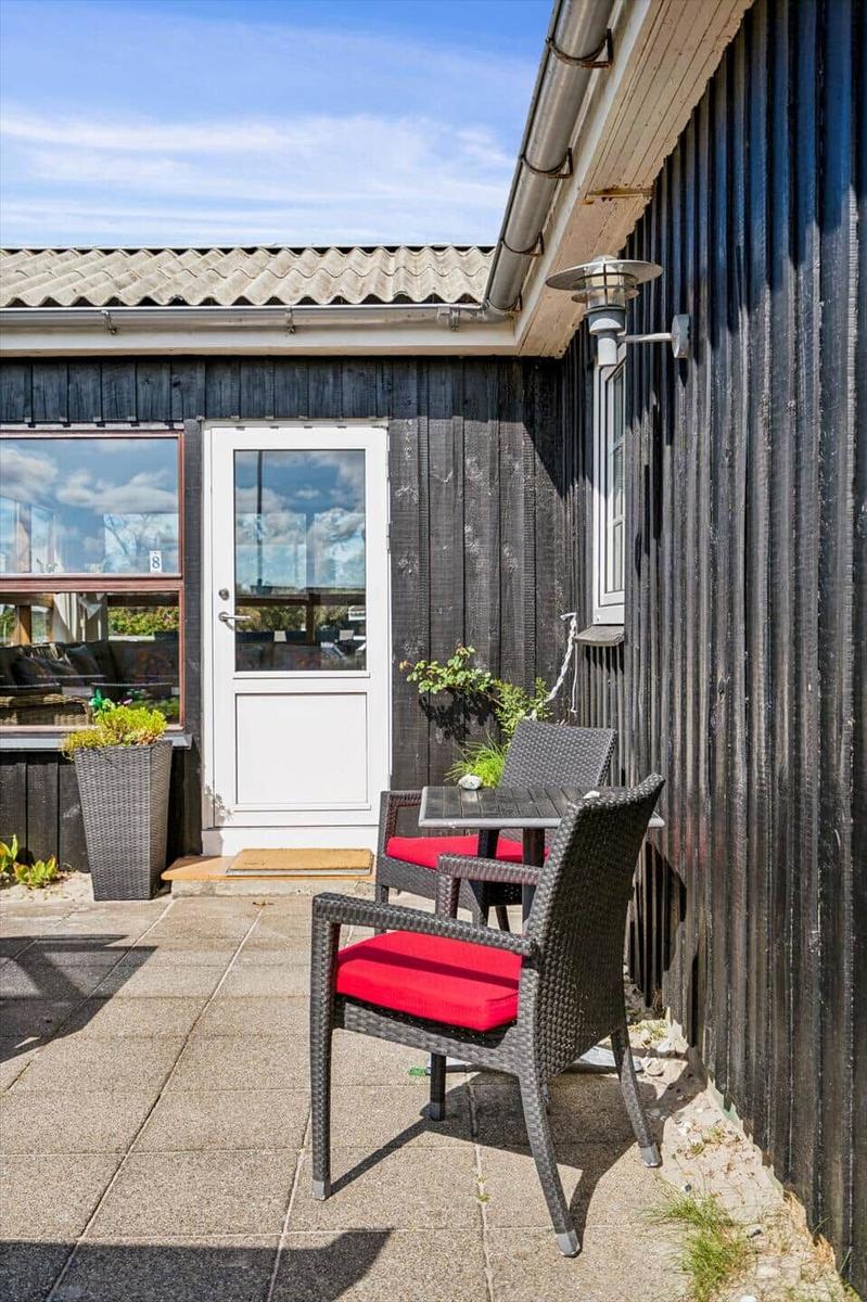 Terrasse mit Tisch und zwei Stühlen mit roten Sitzkissen vor einem Haus mit dunkler Holzfassade.