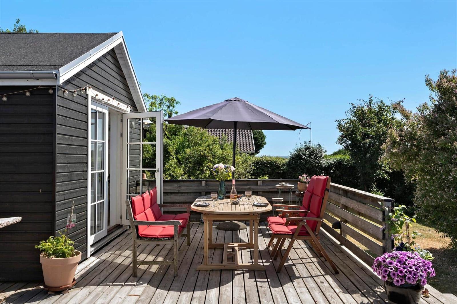 Deck with table, chairs, and umbrella. Wooden floor, plants, and house wall visible.