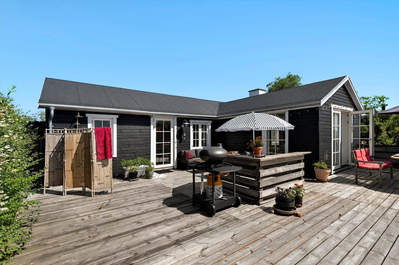 Wooden terrace with grill, seating, and umbrella in front of a black house.