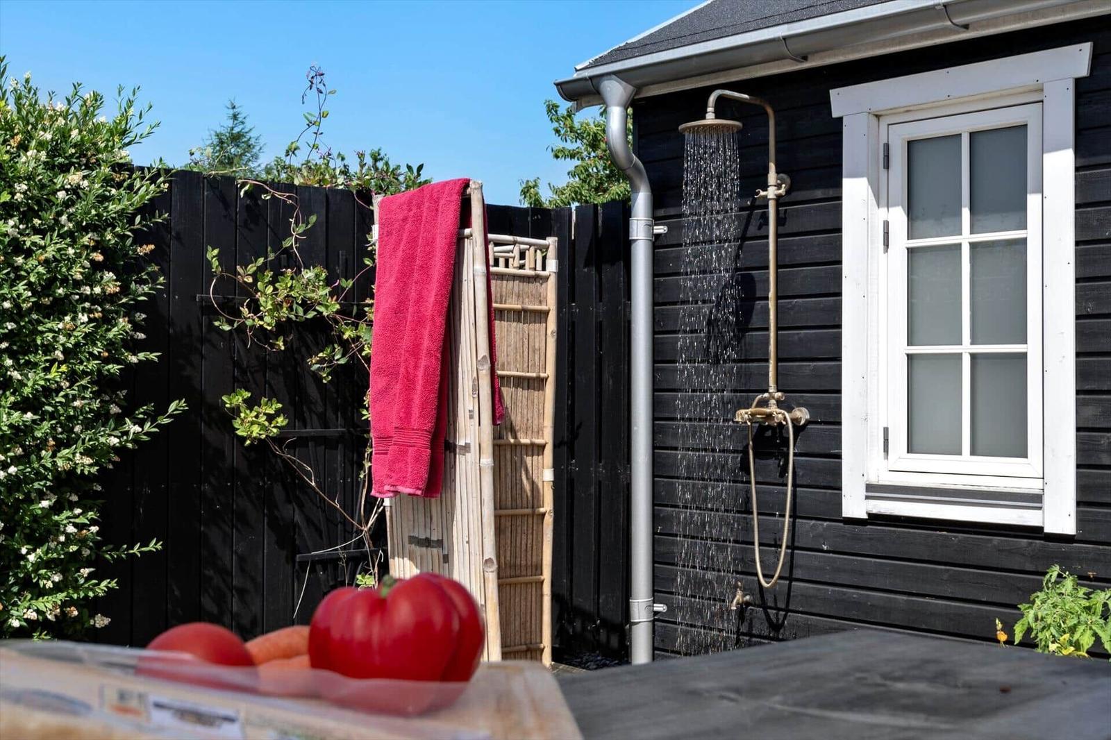 Outdoor shower attached to black house with red towel and tomatoes in foreground.
