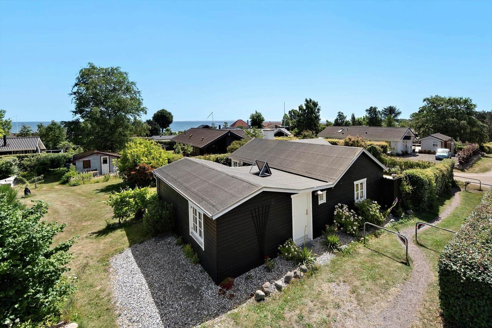 Black wooden house with white door and windows, surrounded by greenery and trees, view of the sea.