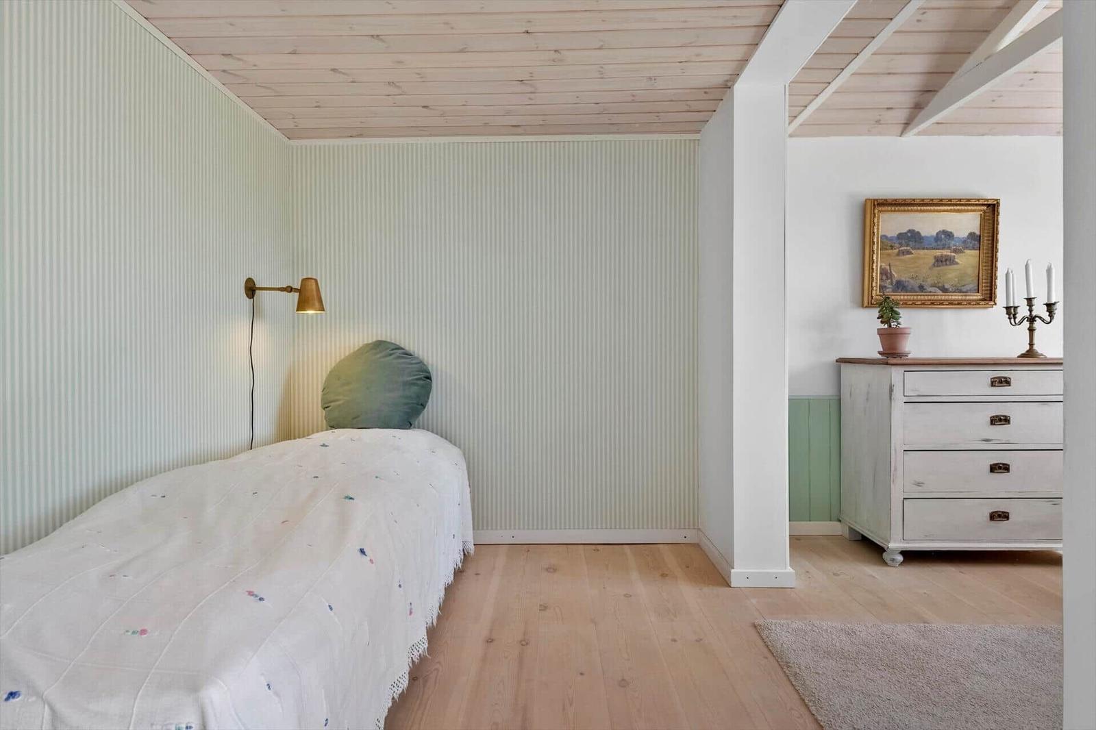 Bedroom with bed, dresser, and wooden floor. Ceiling with beams, wall with striped pattern.
