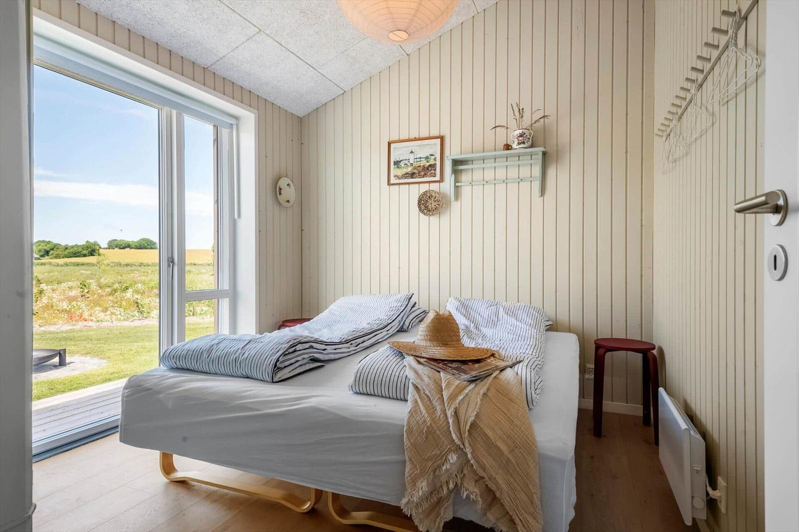Schlafzimmer mit Bett, Fenster und Holzwandverkleidung. Draußen Gras und Bäume.