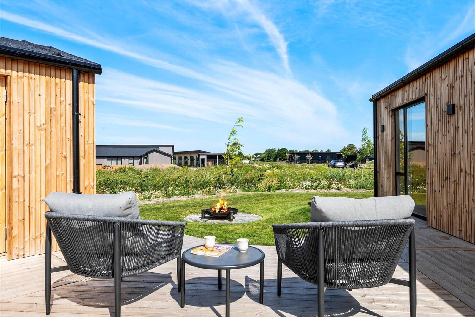 Terrasse mit zwei Stühlen und Tisch, Feuerkorb im Gras, Holzhaus und blauer Himmel.