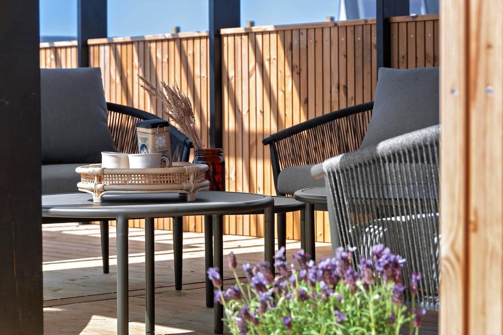 Terrasse mit Sitzgelegenheiten, Tisch und Pflanzen. Holzgeländer und Sonnenschein.