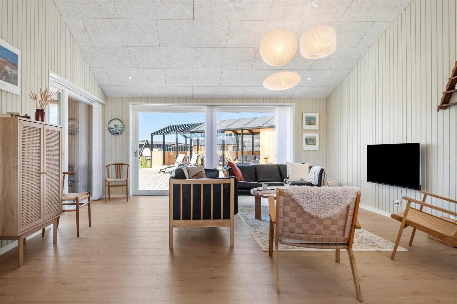 Wohnzimmer mit Holzboden, Sofa, Stühlen und Fernseher. Durchgang zur Terrasse mit Liegestühlen.