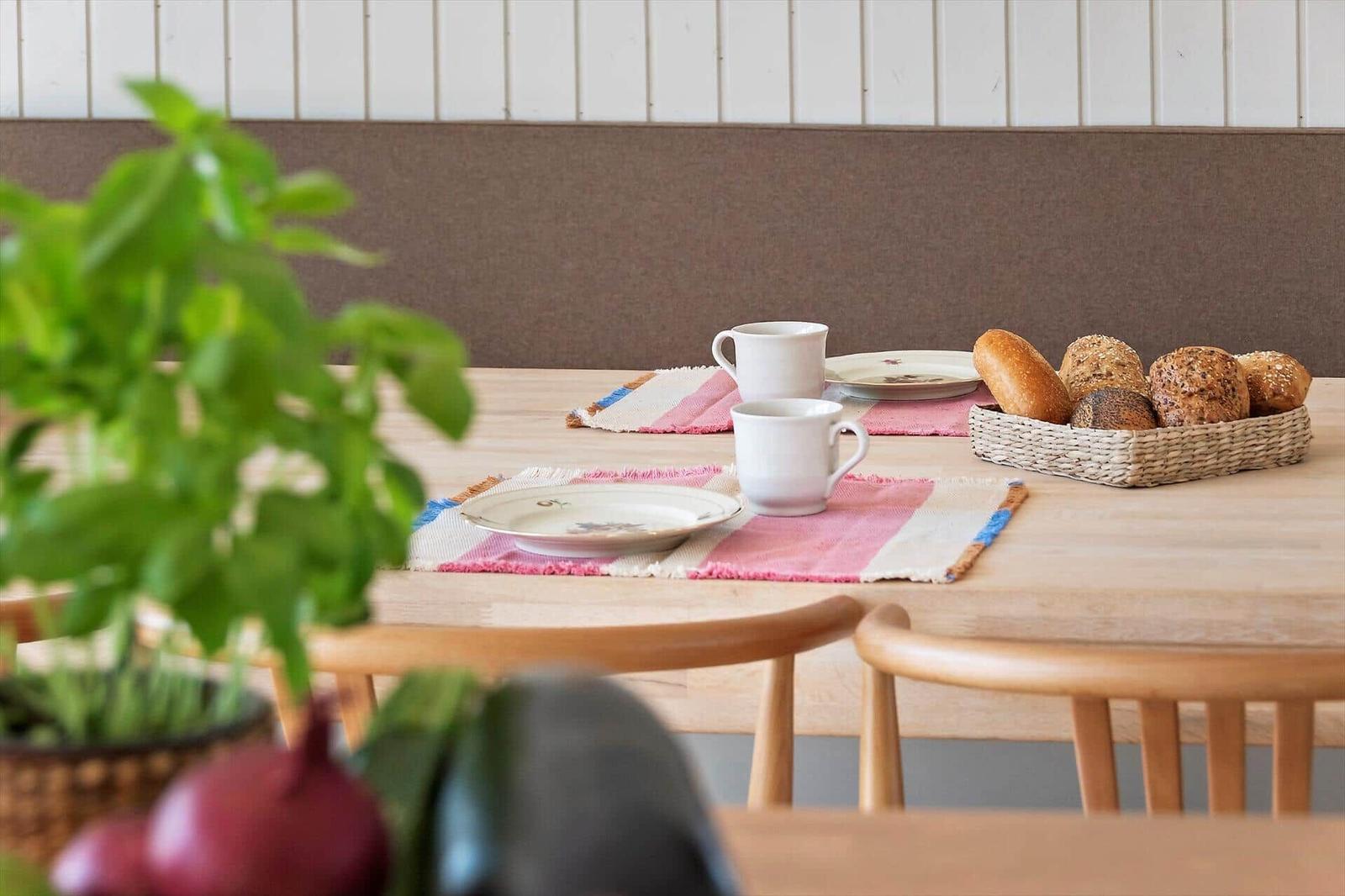 Wooden table with placemats, mugs, plates, and basket of bread.