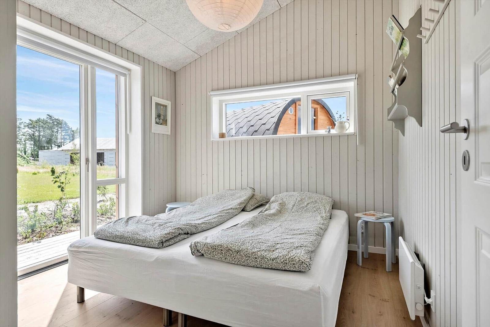 Bedroom with bed, window, and door. Outdoor view with green area and wooden house.