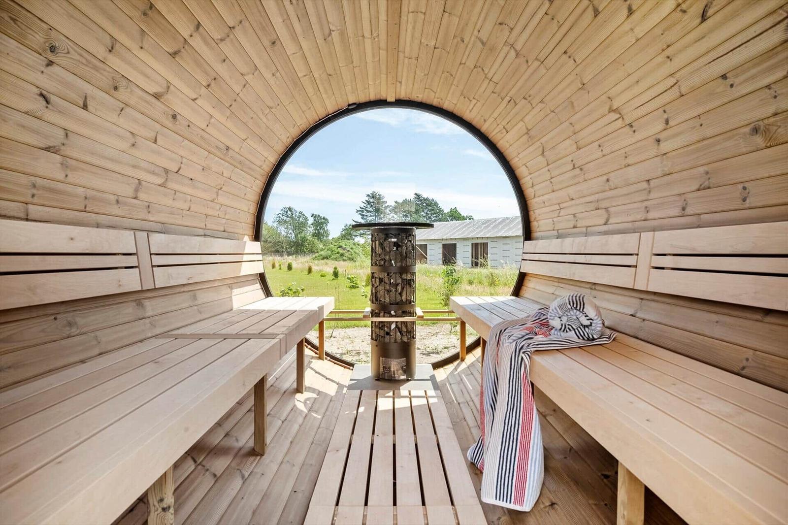 Wood sauna with round window, benches, and wood stove. View of grassland and building.