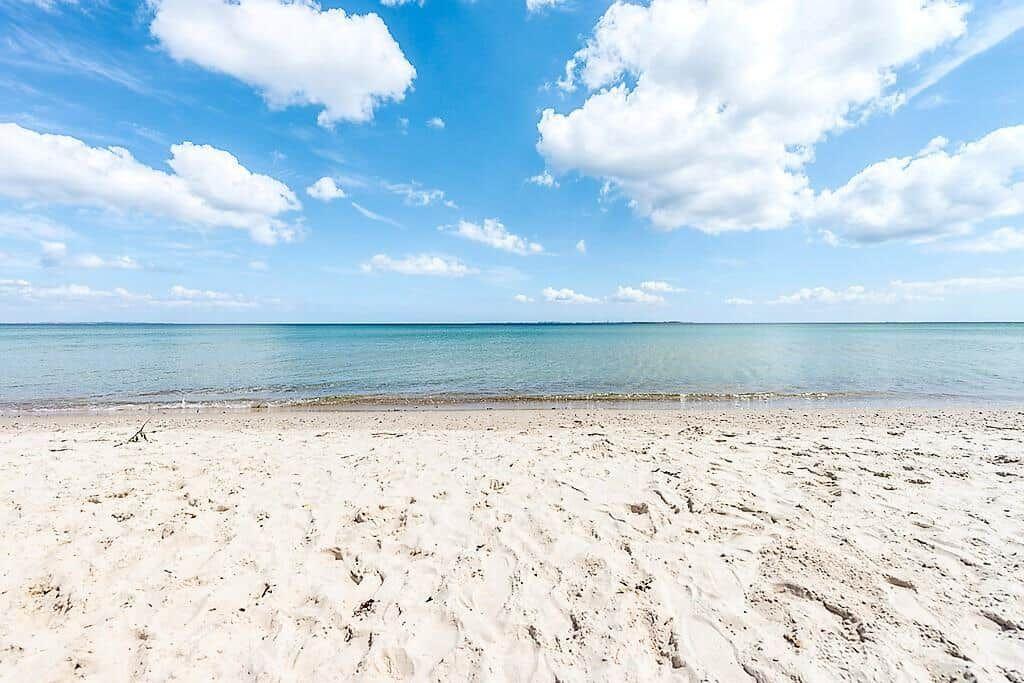 View of the beach with sand and calm sea under blue sky with white clouds.