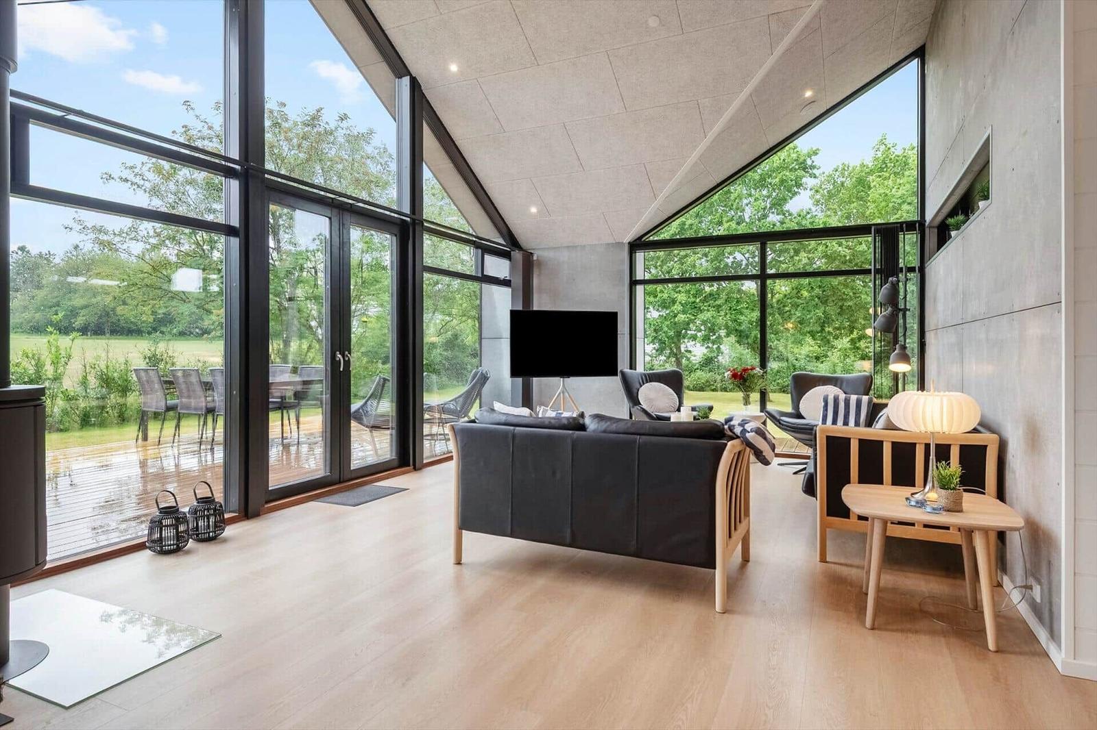 Ein Wohnzimmer mit großem Fenster, Sofa und Fernseher. Draußen ist eine Terrasse mit Tischen und Stühlen.