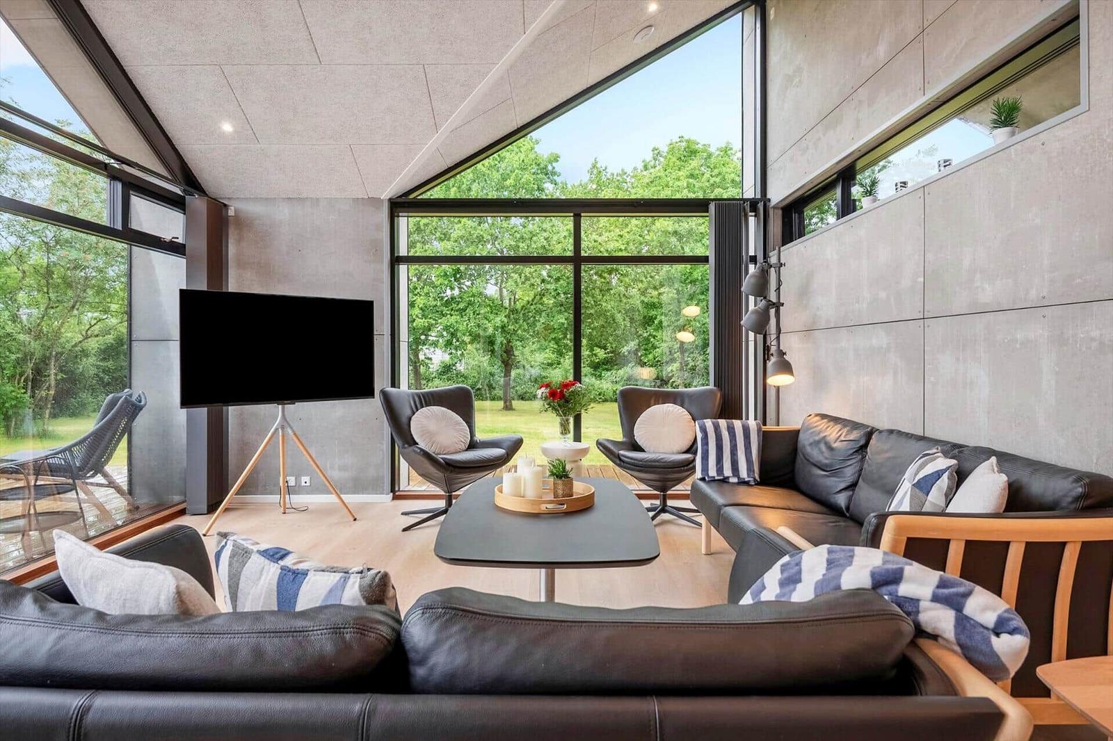 Wohnzimmer mit Sofa, Sesseln und Fernseher. Große Fenster mit Blick auf Grün.