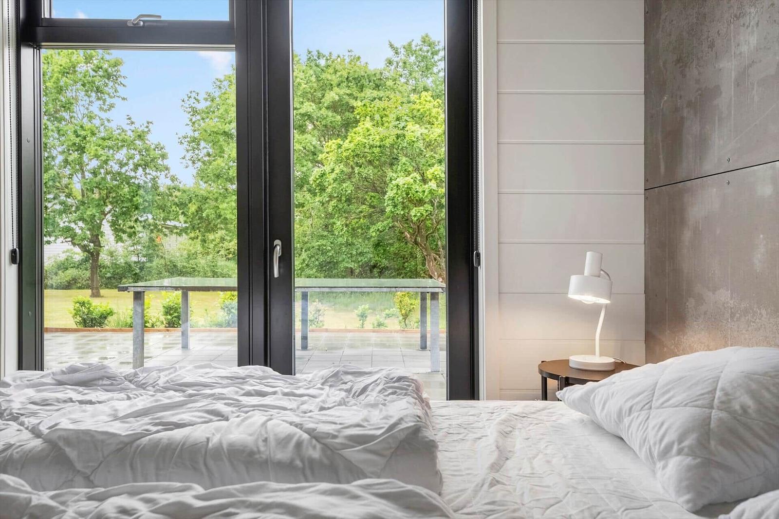 Schlafzimmer mit Blick auf grünen Garten und Terrasse durch große Fenster.