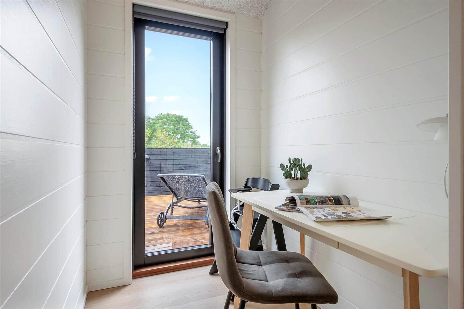 Ein Arbeitszimmer mit Schreibtisch, Stuhl und Pflanze. Durch die Glastür ist eine Terrasse mit Liegestuhl sichtbar.
