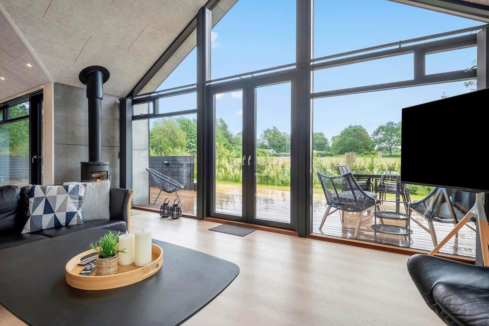 Wohnzimmer mit Kamin, großem Fenster und Außenterrasse mit Sitzgelegenheiten.