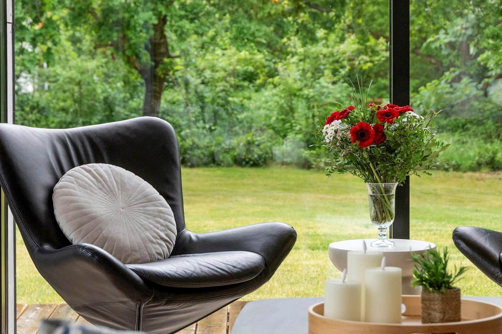 Schwarze Ledersessel mit Kissen, Blumenstrauß und Kerzen vor großem Fenster mit Blick auf Grün.