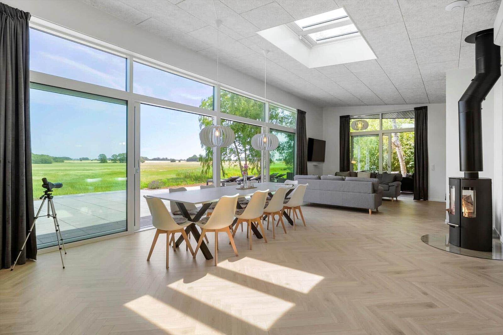 Großes Wohn- und Esszimmer mit bodentiefen Fenstern und Holzboden. Holzofen und Fernseher sichtbar.