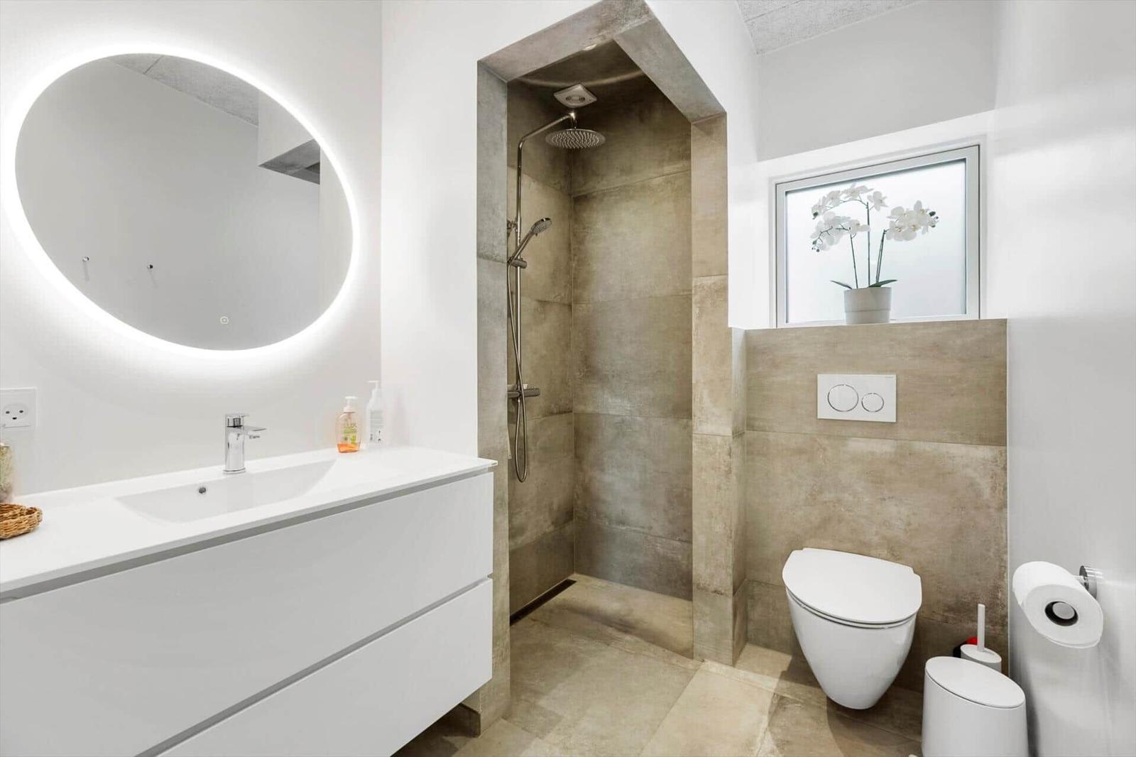Moderne Badezimmer mit Dusche, Waschbecken und Toilette. Runder beleuchteter Spiegel und Fenster mit Orchidee.