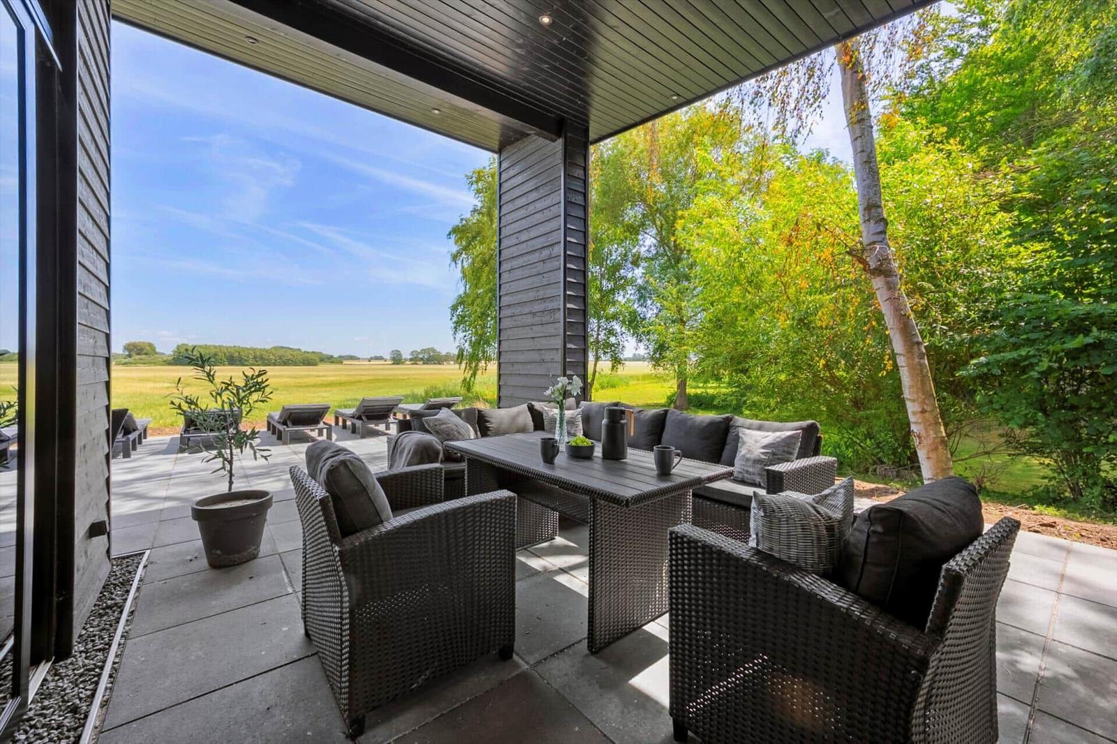 Terrasse mit Sitzgruppe und Tisch unter Dach, Blick auf Feld und Bäume.
