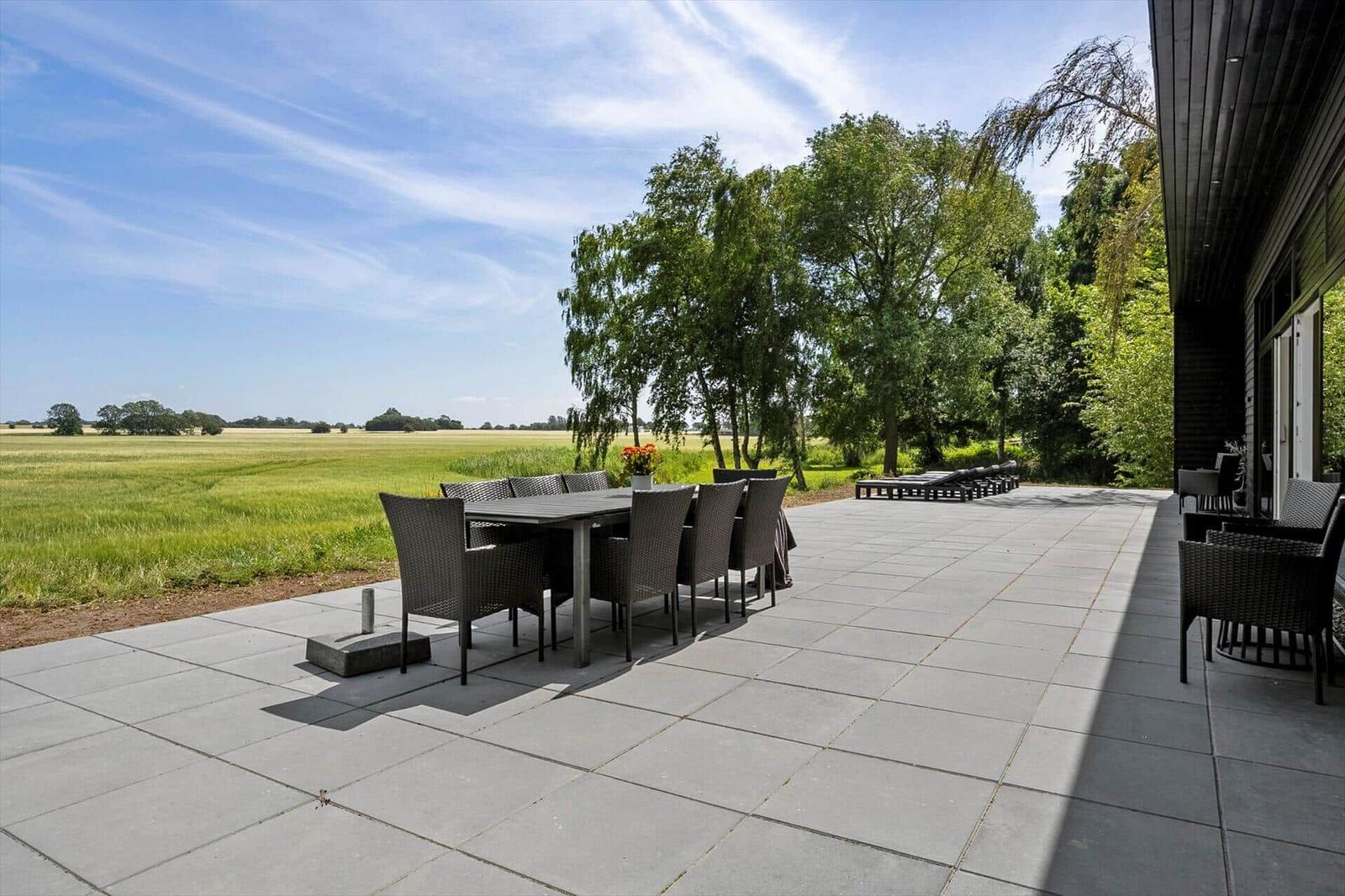 Großer Terrassenbereich mit Tisch, Stühlen und Blick auf Felder und Bäume.