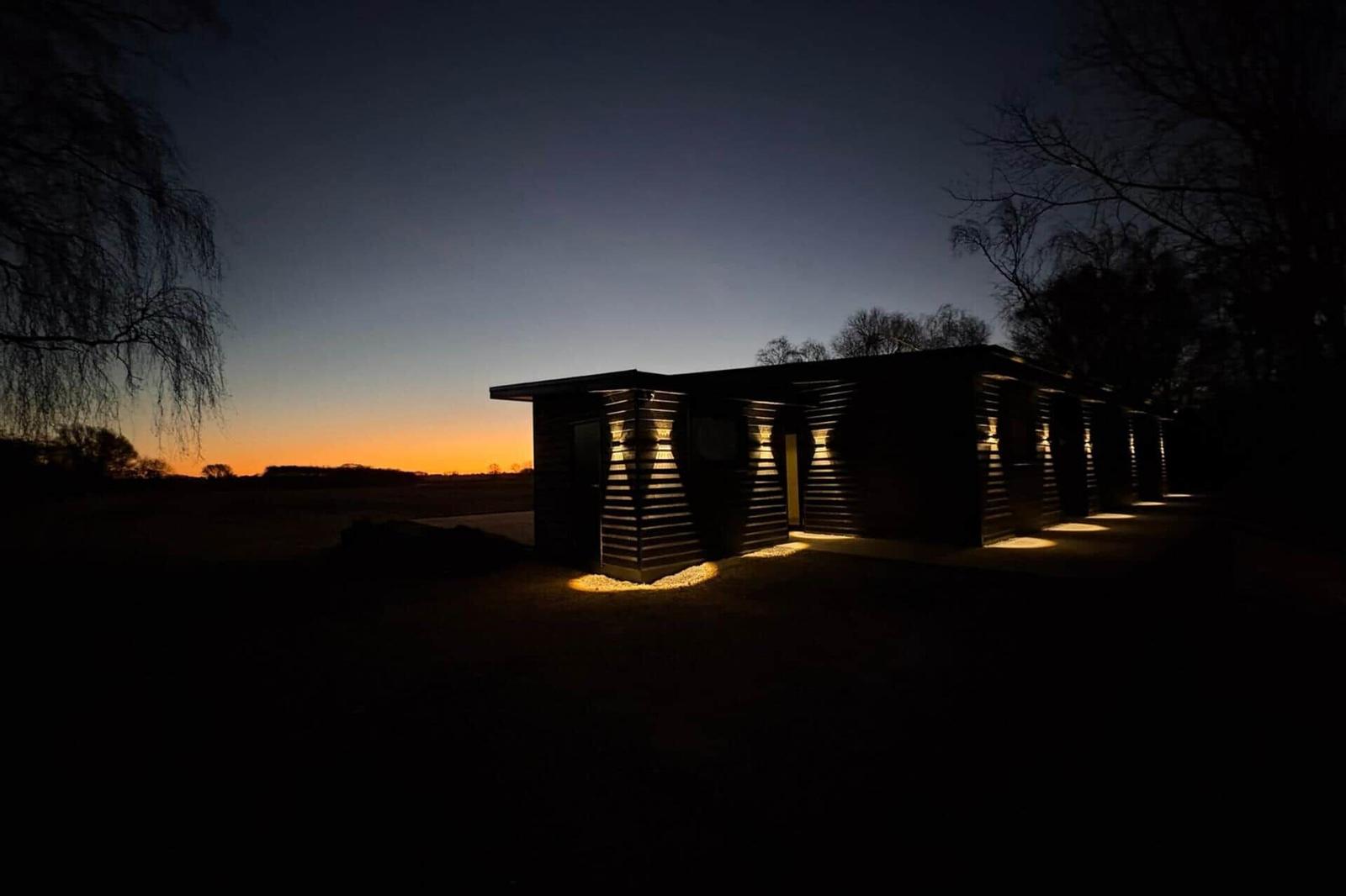 Ein modernes Holzhaus bei Dämmerung mit Außenbeleuchtung und Blick auf Felder.