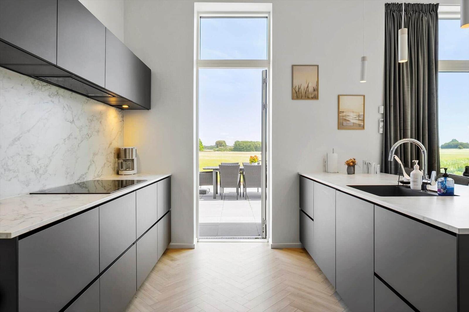 Moderne Küche mit Blick auf Terrasse und Feld. Weiße Arbeitsplatten und graue Schränke.