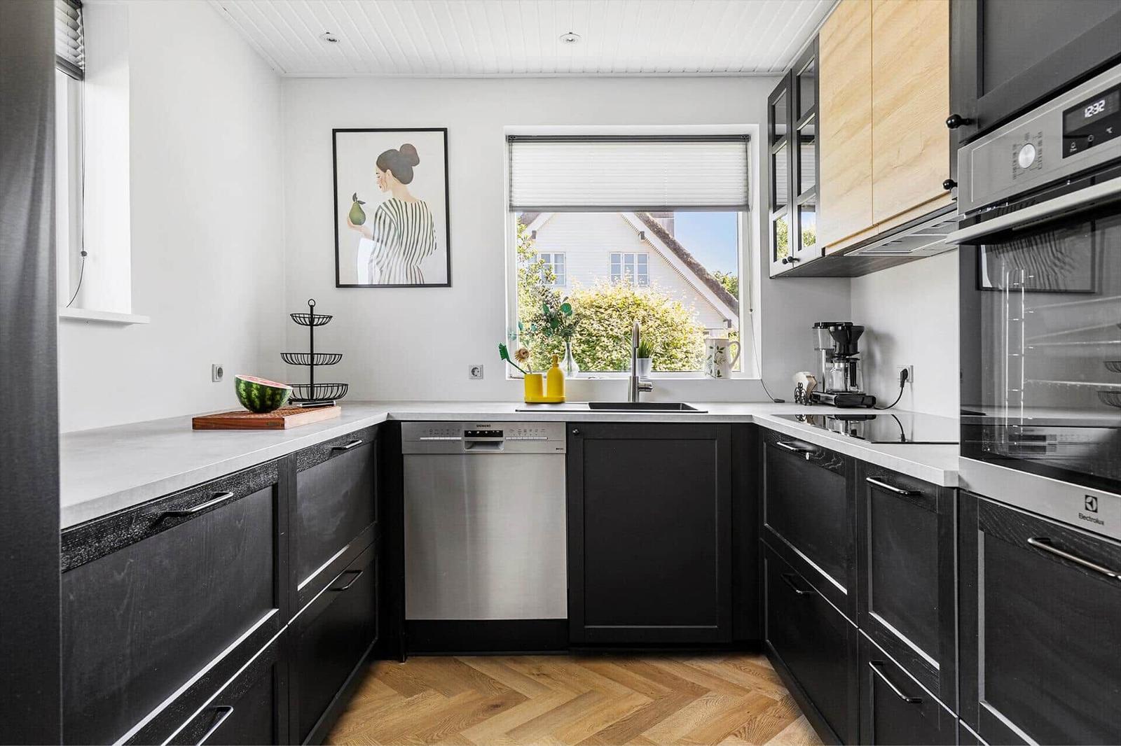 Modern kitchen with black cabinets, wooden floor, and window view of house and greenery.