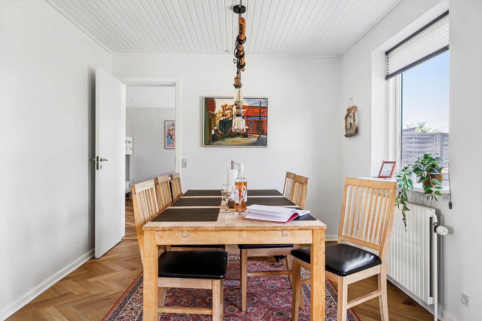 Dining room with wooden table, chairs, and window. Wall painting and ceiling light visible.