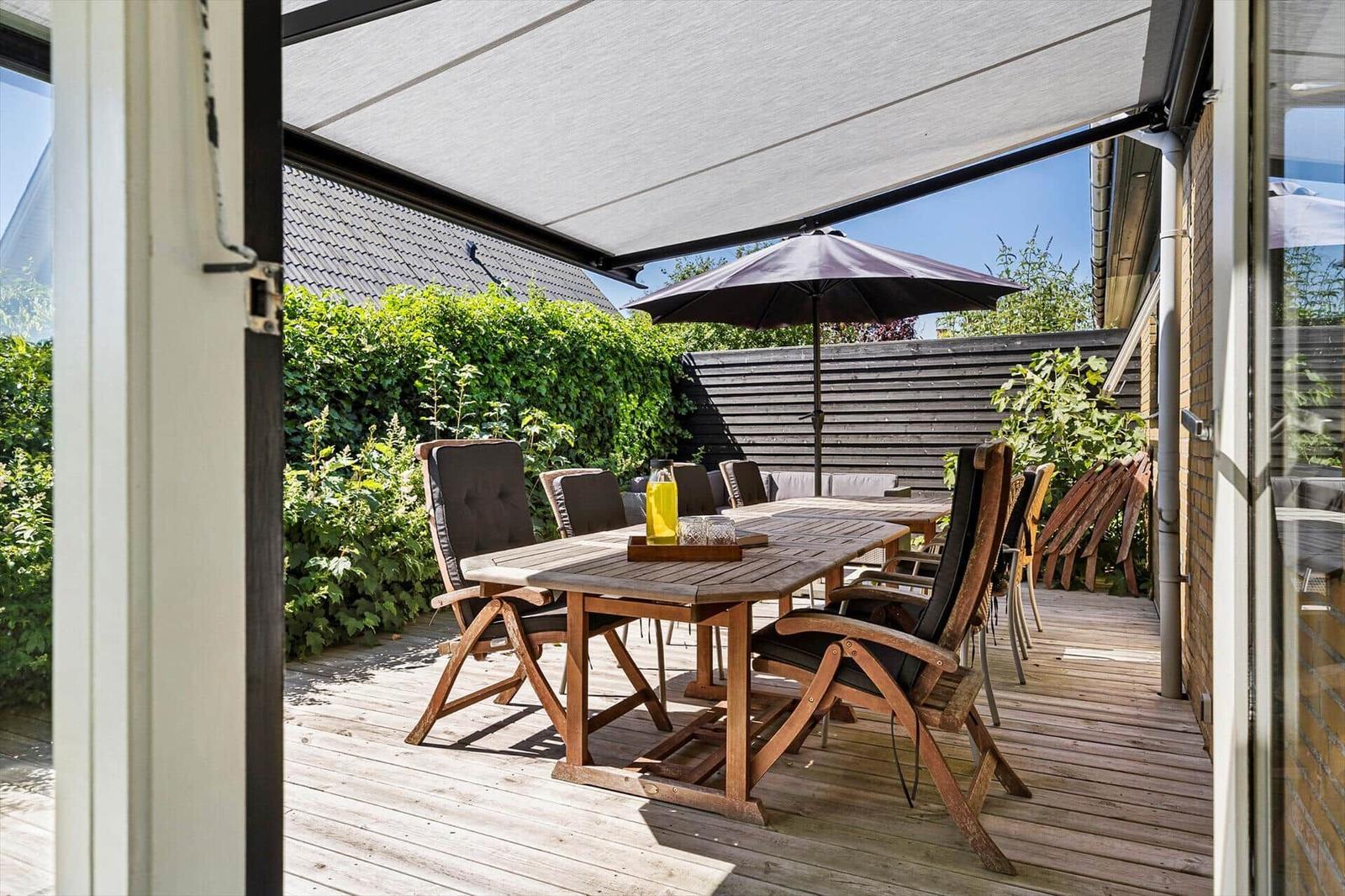 Terrace with wooden table, chairs, and sunshade. Background: green hedge and wooden house wall.