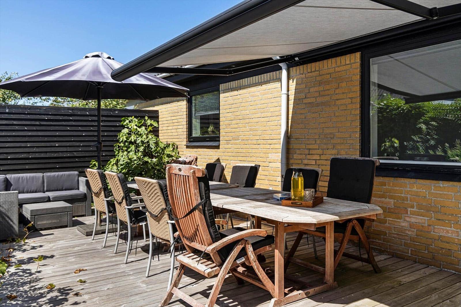 Deck with wooden table, chairs, and awning. Background: yellow brick wall and garden.