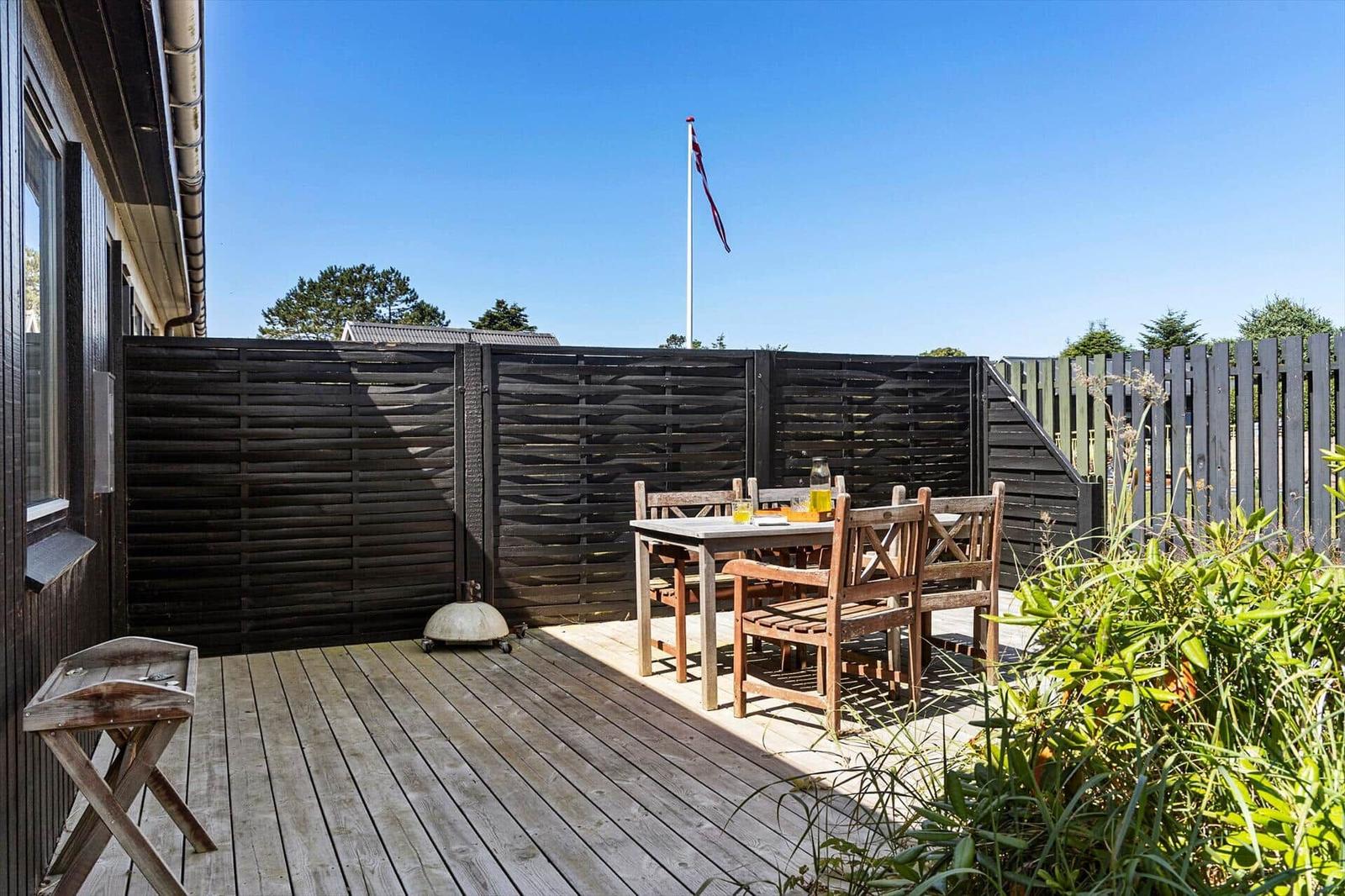 Wooden terrace with table, chairs, and grill. Background: fence and flag under blue sky.