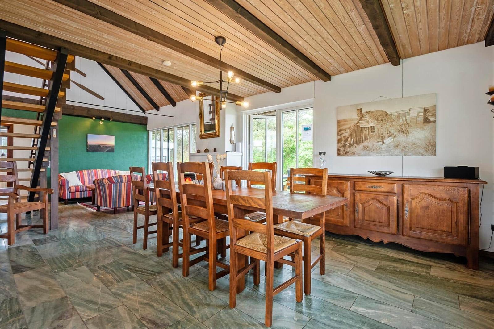Esszimmer mit Holztisch, Stühlen und Holzdecke. Nebenraum mit Sofa und Treppenhaus.