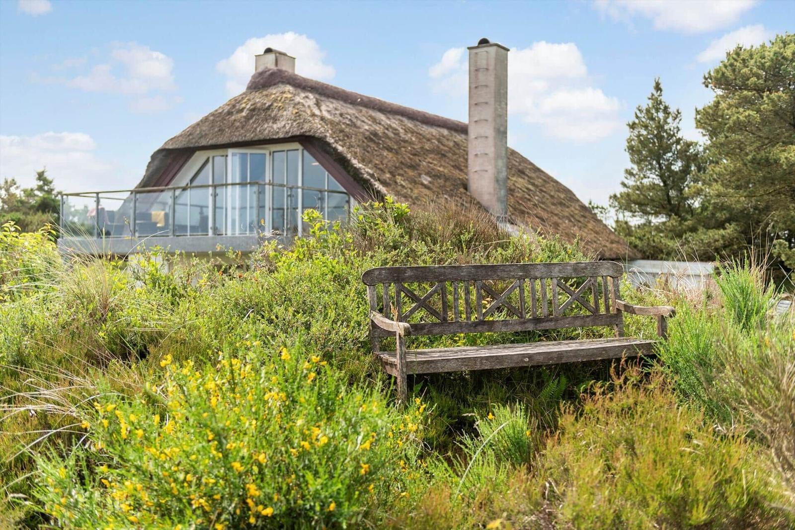 Haus mit Strohdach, Holzbank im Garten, Glasbalcony, umgeben von Gräsern und Bäumen.