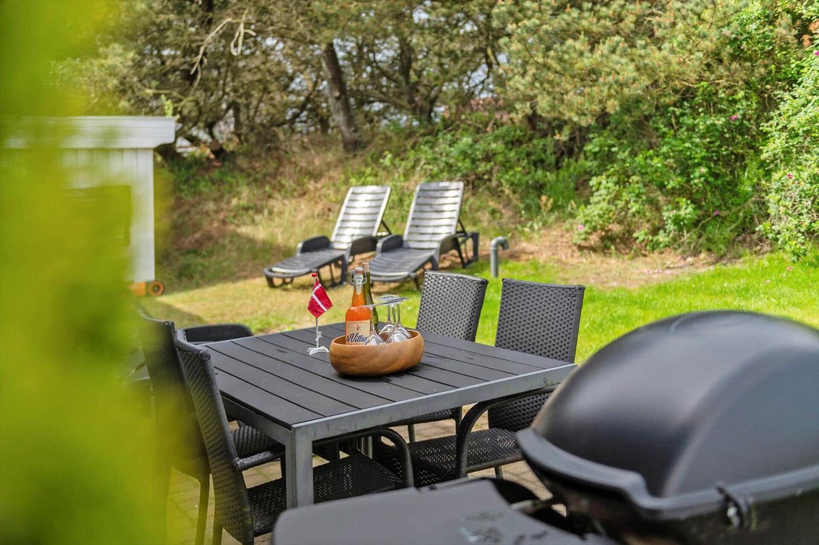 Außenbereich mit Tisch, Stühlen, Liegen und Grill. Eine dänische Flagge und Flaschen auf dem Tisch.