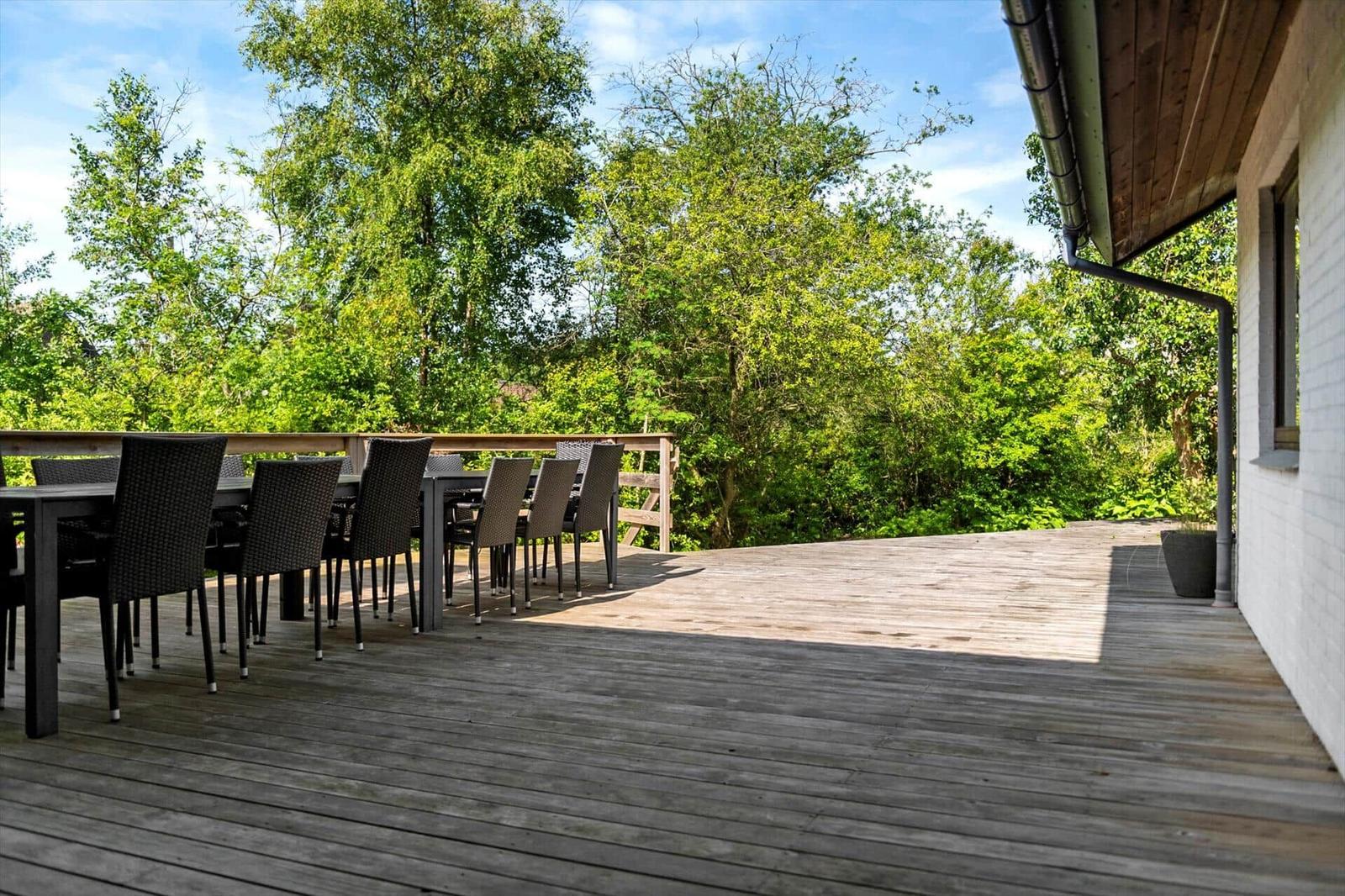Holzterrasse mit Tisch und Stühlen, umgeben von grüner Vegetation und einem Haus.