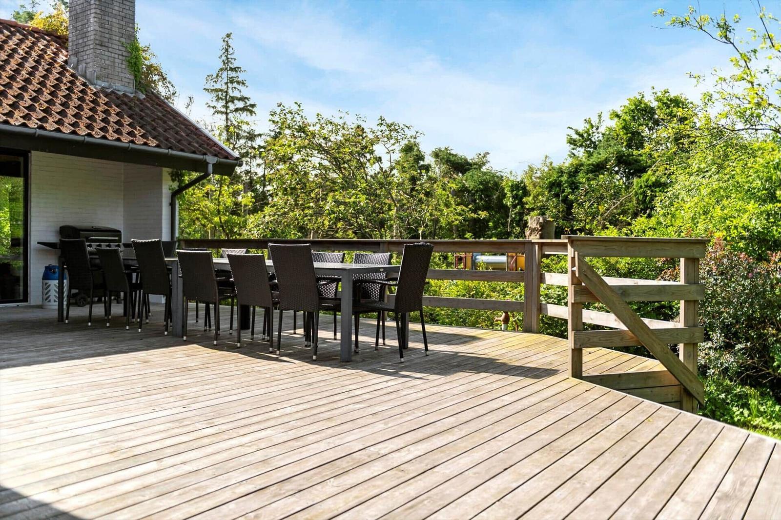 Holzterrasse mit Sitzgruppe und Grill, umgeben von Bäumen und einem Haus.