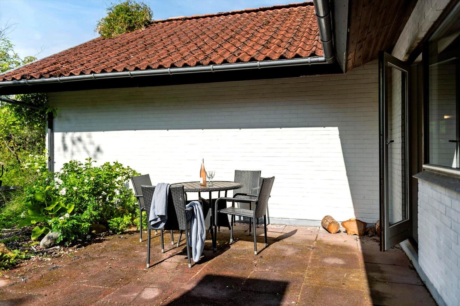 Terrasse mit Tisch und Stühlen unter Dach. Weiße Wand, rote Ziegeldach, Gartenbereich.