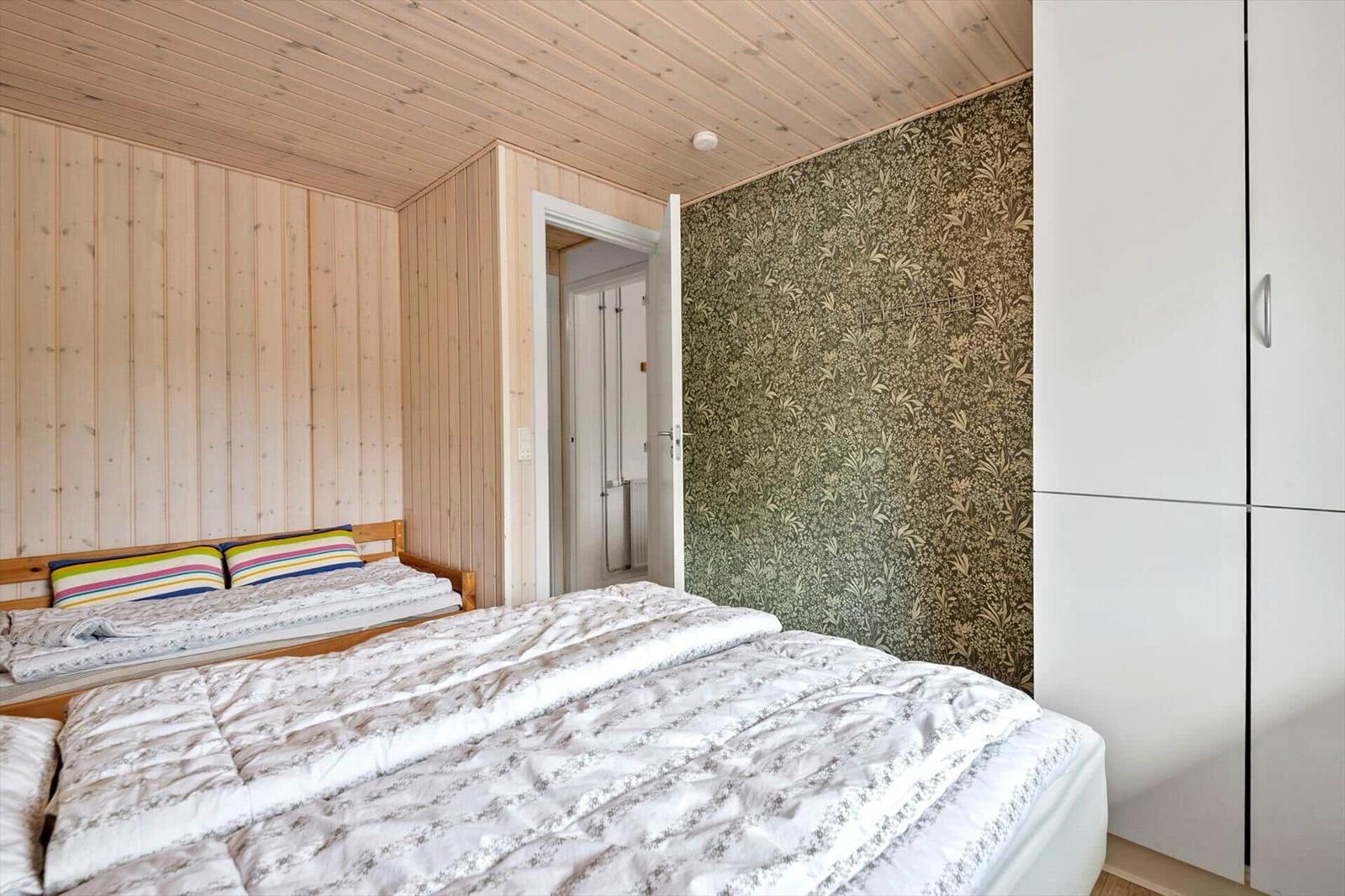 Schlafzimmer mit Doppelbett, Holzwand und gemustertem Wandbezug. Weiße Schrankwand und Tür sichtbar.