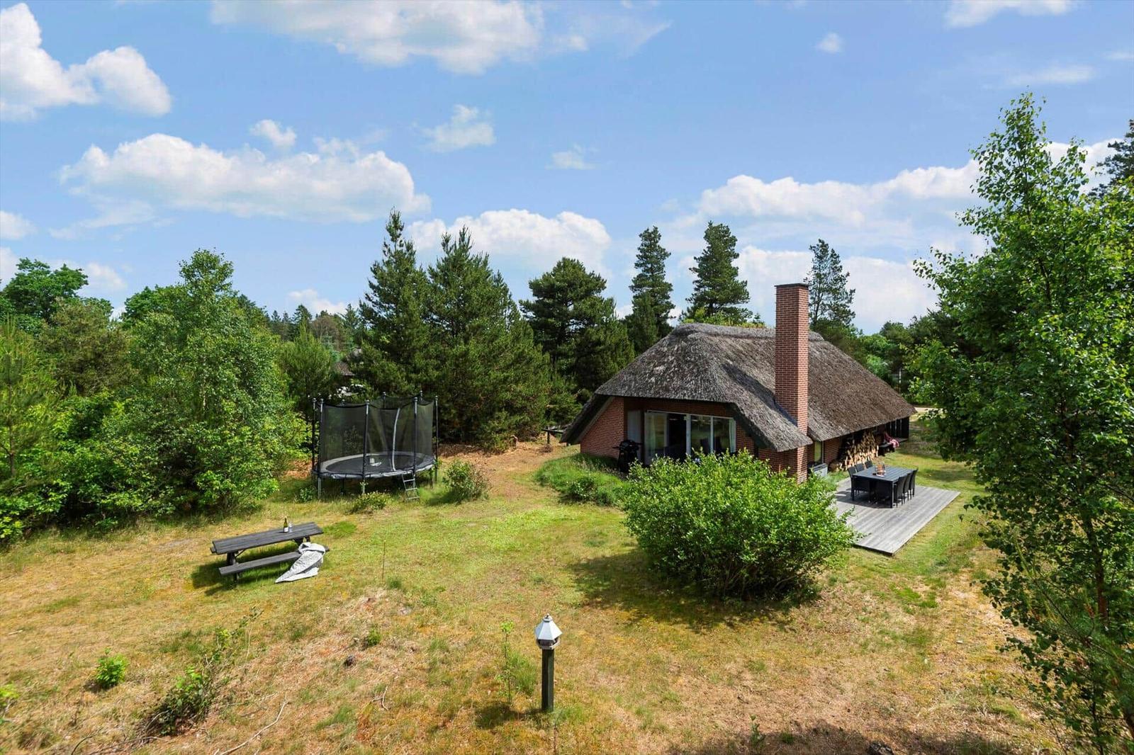 Haus mit strohgedecktem Dach, Terrasse, Tisch, Trampolin und Grillplatz im Grün.