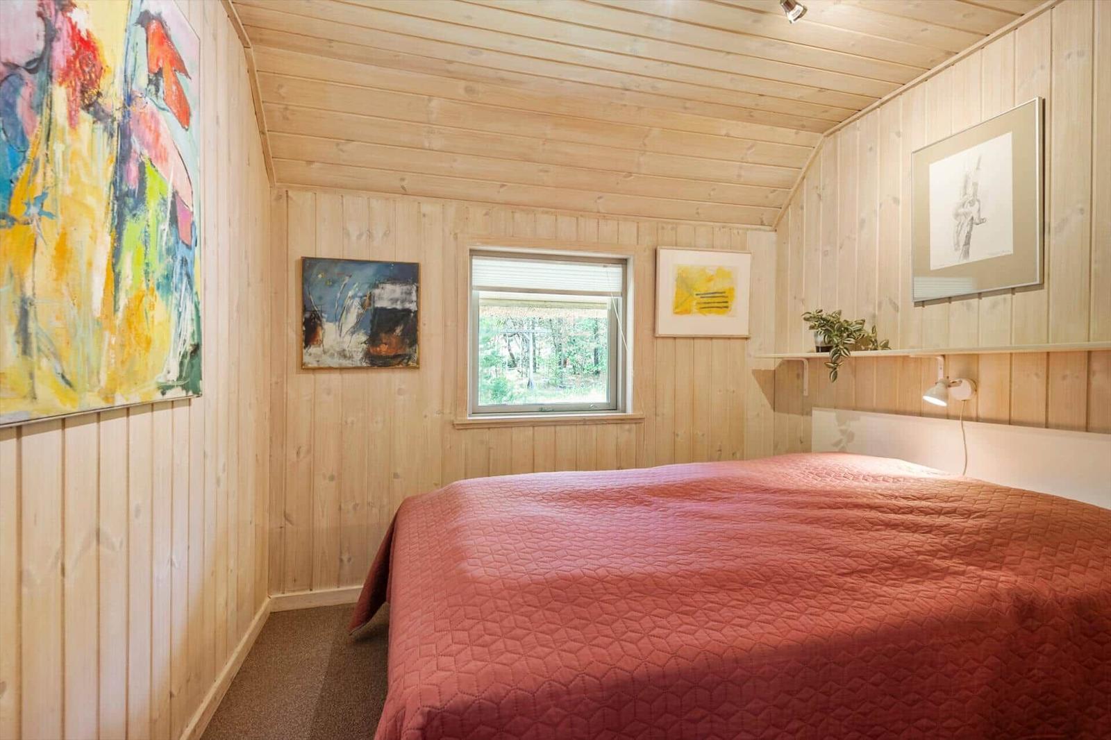 Schlafzimmer mit Holzwänden, Bett mit roter Decke und Fenster mit Blick auf Wald.