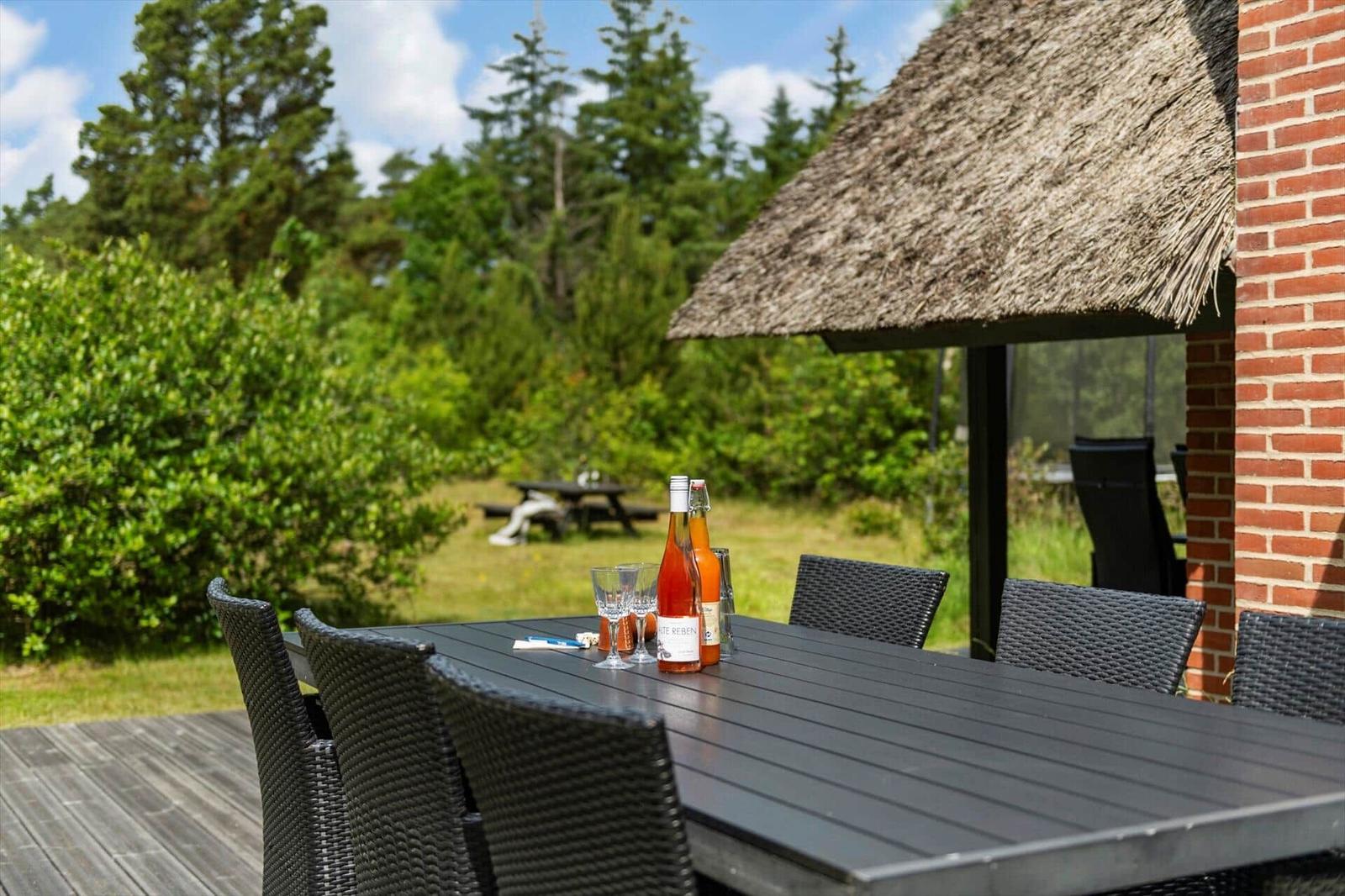 Außenbereich mit Tisch und Stühlen unter einem Strohdach. Im Hintergrund sind Bäume und ein Picknicktisch zu sehen.