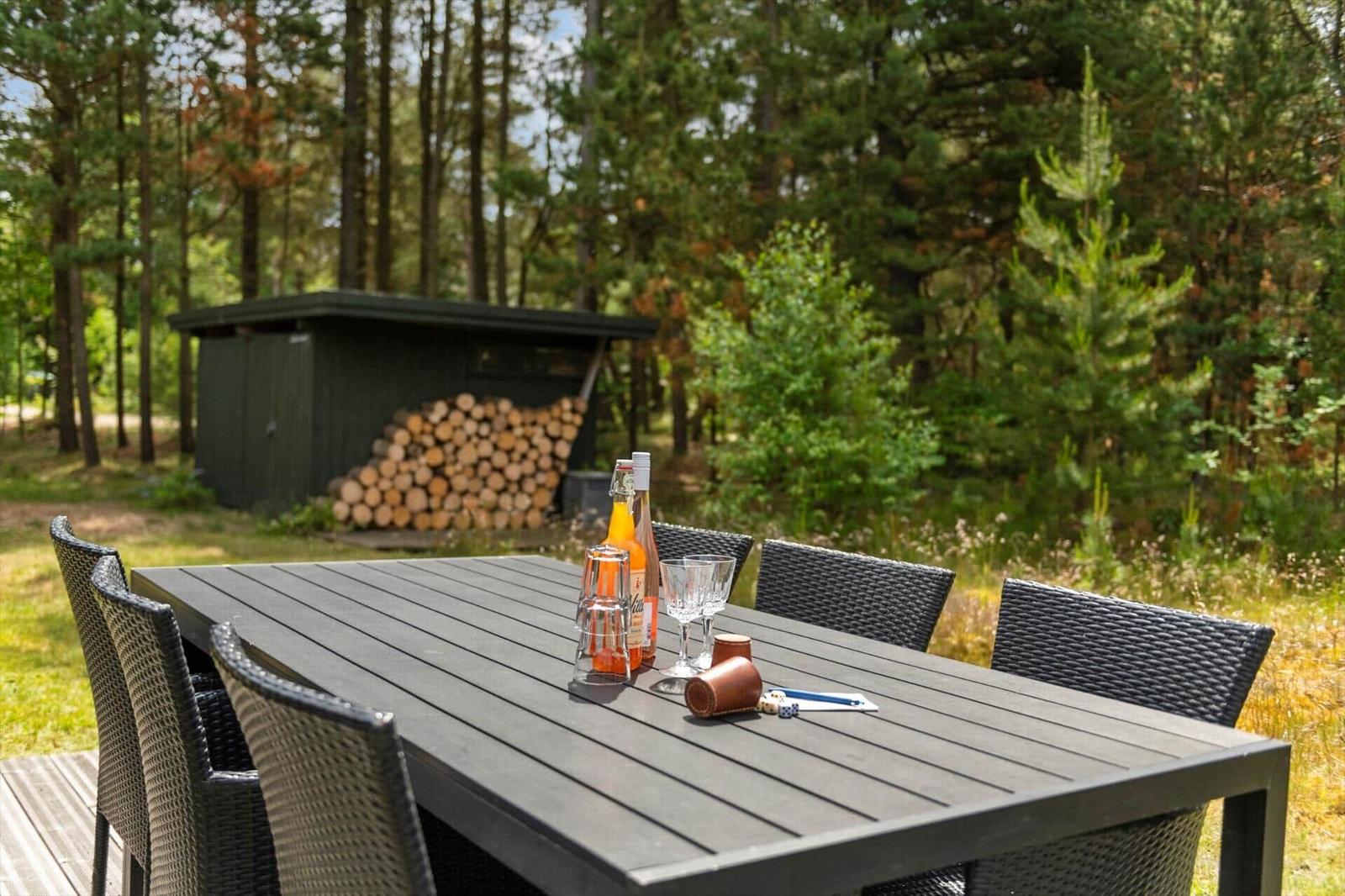 Hochwertiger Holztisch mit Stühlen im Wald. Hintergrund: Holzstapel und kleiner Schuppen.